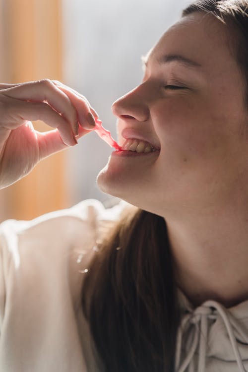 Free Happy young crop female with eyes closed smiling and biting sweet marmalade on blurred background Stock Photo