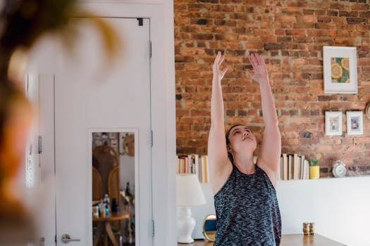 A woman practicing yoga with hands up indoors against a brick wall.