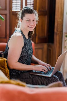A woman smiling while typing on a laptop in a cozy home setting.