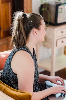 A woman types on a laptop, working remotely in a cozy home setting with natural light.
