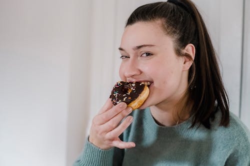 Free Delighted young female biting delicious sweet chocolate donut and looking at camera while smiling Stock Photo