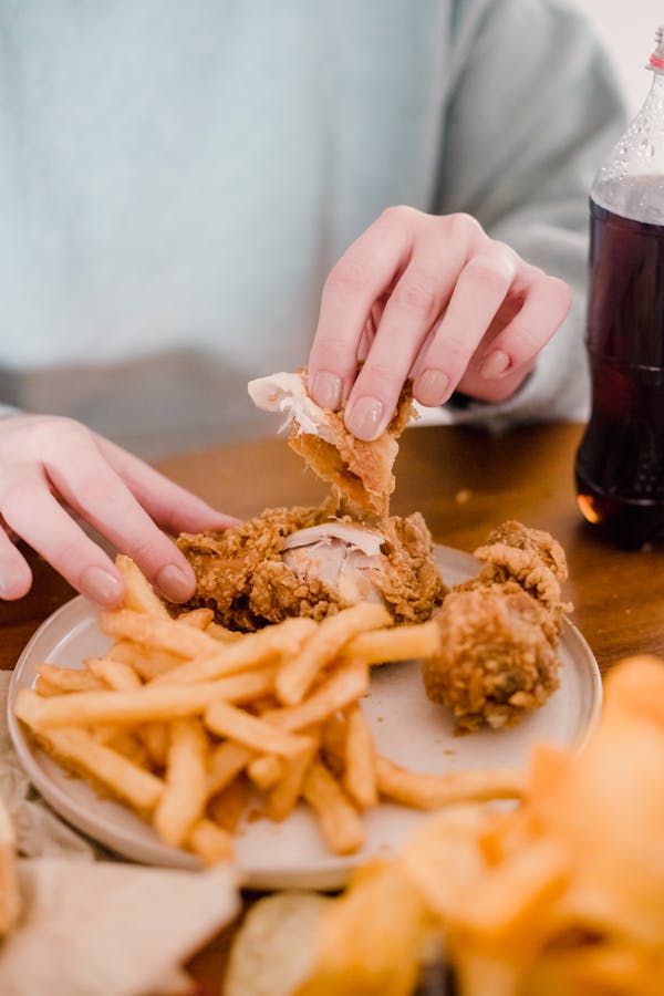 Crop anonymous female eating fried chicken in breading with fries at table with lemonade