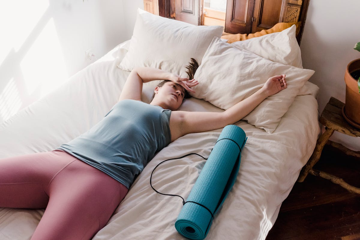 High angle of exhausted female in sportswear lying on bed with hands above head near sports mat after training
