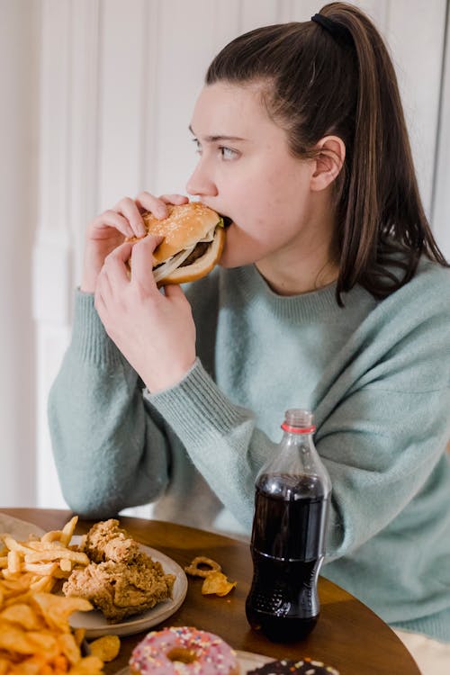 Serious female biting delicious burger while sitting at table with various junk food and soda in light room at home