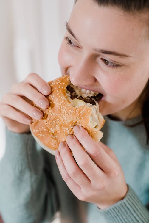 Free Crop female in casual wear biting tasty hamburger with fresh bread while having lunch with junk food in light room at home Stock Photo