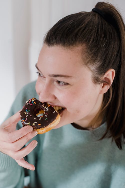 Free From above of happy young female with dark hair eating yummy doughnut and smiling while looking away Stock Photo