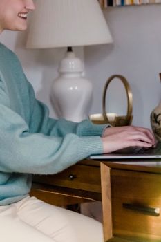 Side view of crop positive female sitting near desk and working on laptop in bedroom