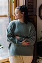 Thoughtful female with folded arms wearing knitted pullover and dreamy contemplating while standing near window