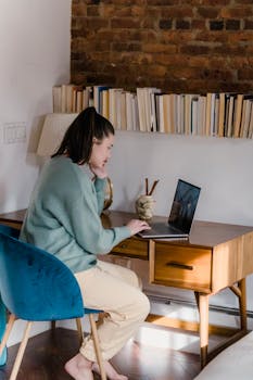 Woman in cozy home office working on a laptop, surrounded by books and warm decor.