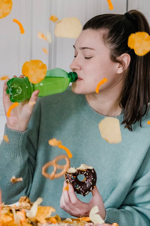 Free Delighted female with closed eyes showered by chips and crisps drinking fresh cold soda and eating yummy donut near heap of yellow snacks Stock Photo
