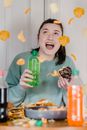 Cheerful young lady with opened mouth holding glazed chocolate bitten donut and bright bottle with fresh drink and surprising at shower of crisps and chips