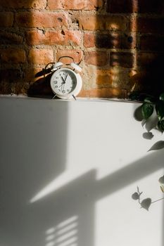Composition of white alarm clock under sunlight from window illuminating brick wall and fridge with shadows from shutters