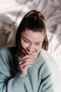 From above of charming female in blue sweater putting hair tip in mouth and smiling while sitting in bedroom under daylight and looking away