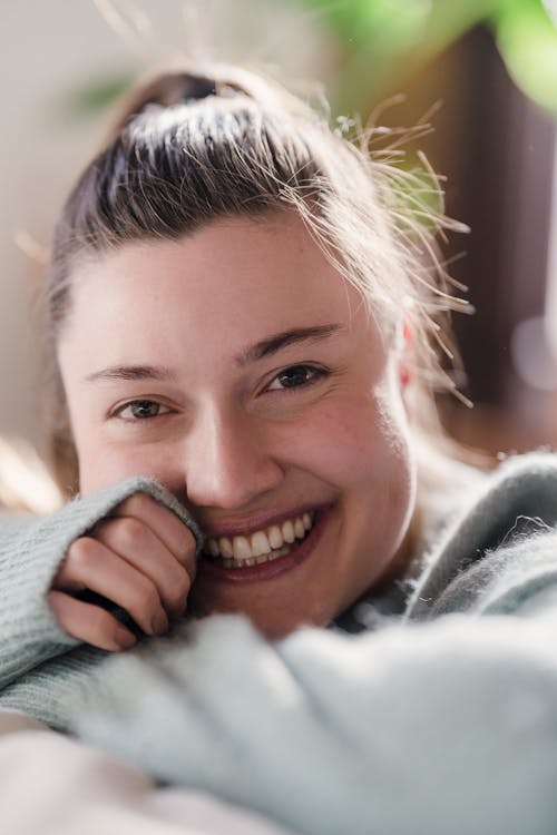 Portrait of cheerful young brunette putting head on arms folded on bed and looking at camera with open smile