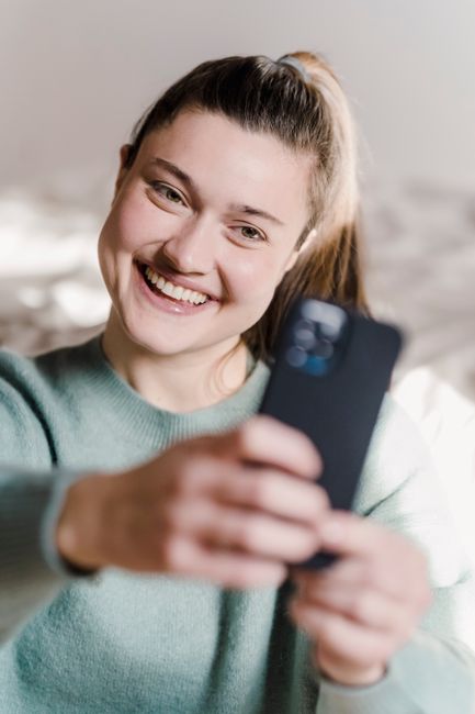 Happy smiling female with ponytail and sincere toothy smile looking at smartphone screen while taking self shot in sunny room