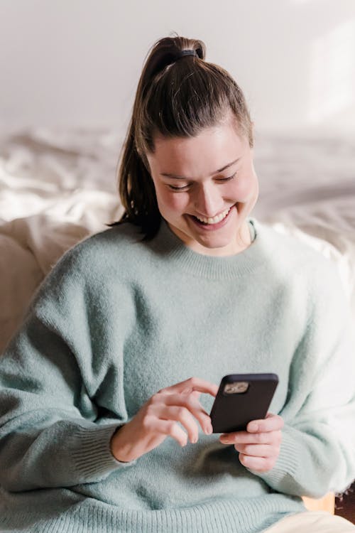 Free Happy smiling female in warm pullover sitting near bed and looking down while resting in apartment and browsing smartphone Stock Photo
