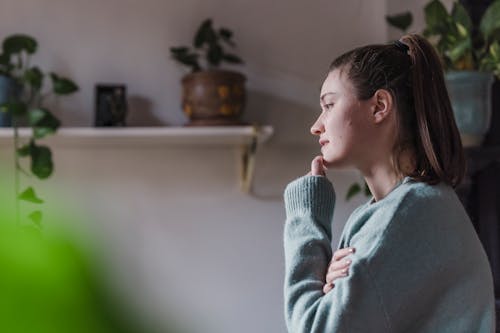 Side view of young pensive female in room with shelves with potted plants touching face while pondering