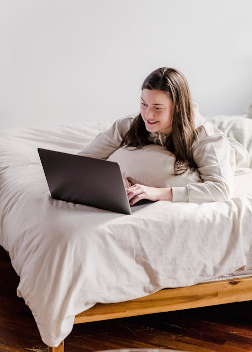 Happy young female millennial with long hair in casual clothes smiling while lying on comfortable bed and watching funny video during weekend at home