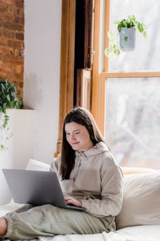 Smiling young female freelancer in casual clothes using laptop while sitting on sofa and working remotely from home