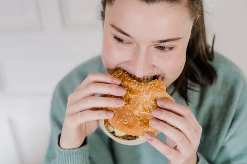 Free From above of crop female biting appetizing hamburger in hands and looking away on blurred background Stock Photo