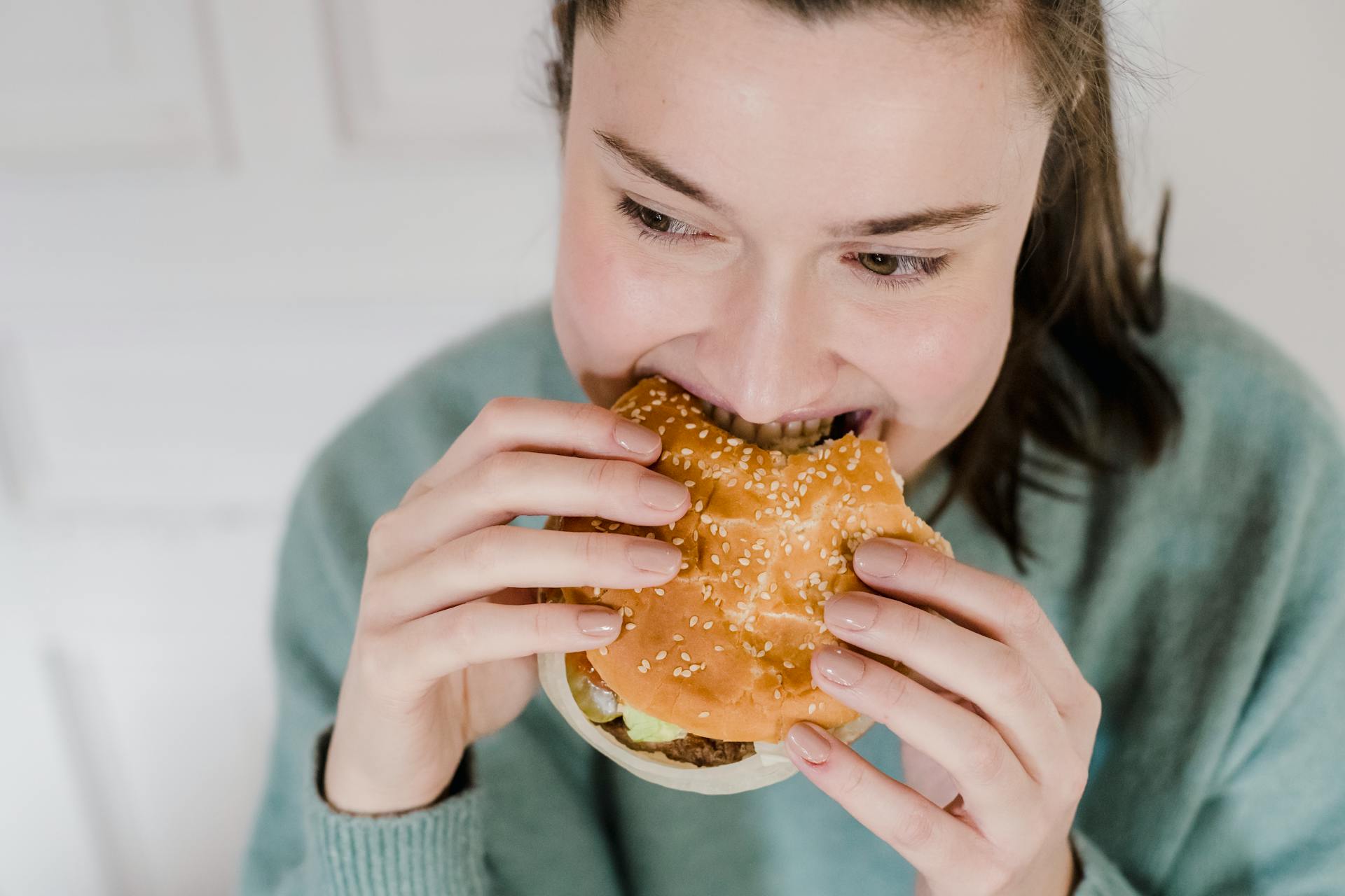 From above of crop female biting appetizing hamburger in hands and looking away on blurred background