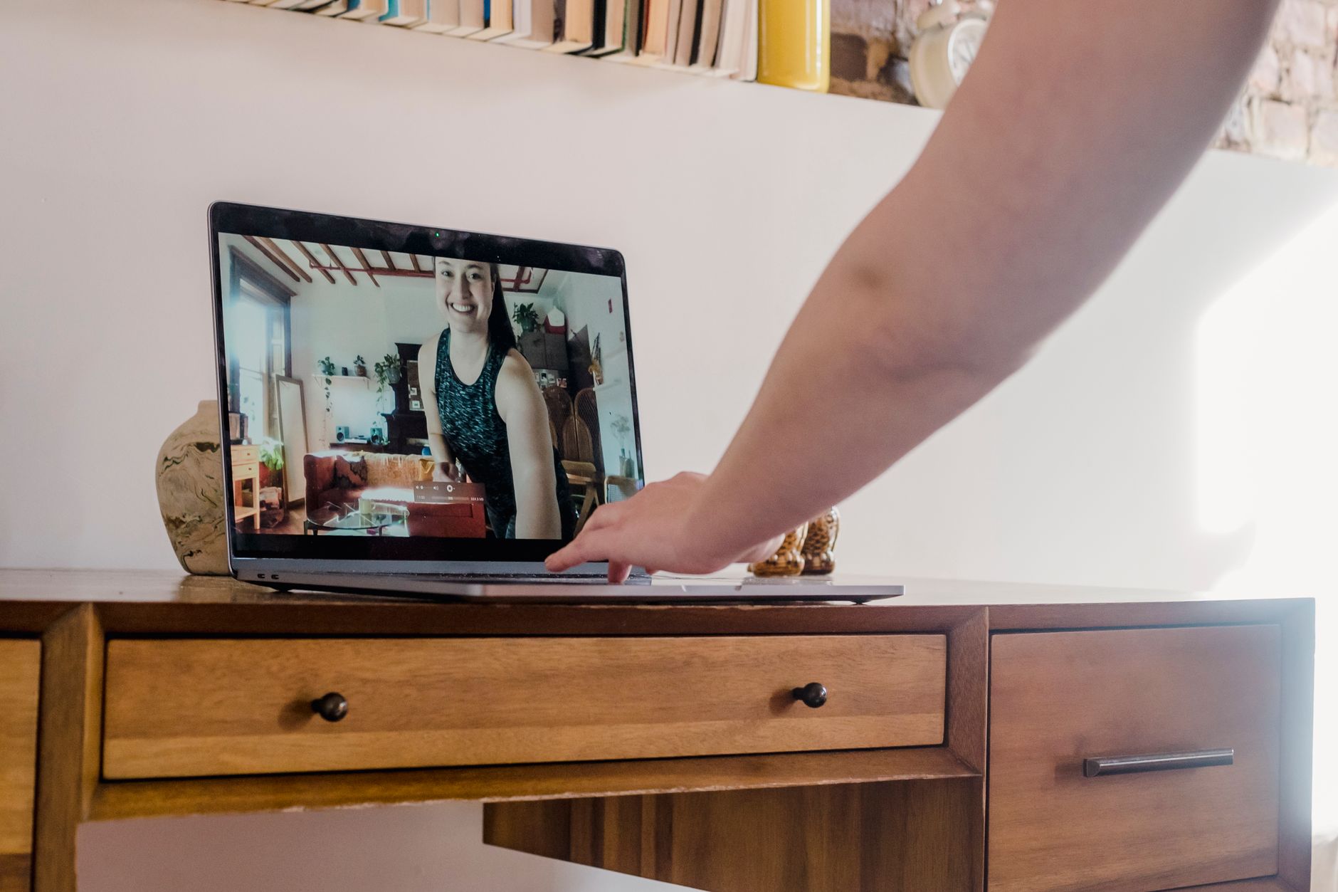 Hand opening a laptop on a wooden desk, screen showing a smiling woman on a video call