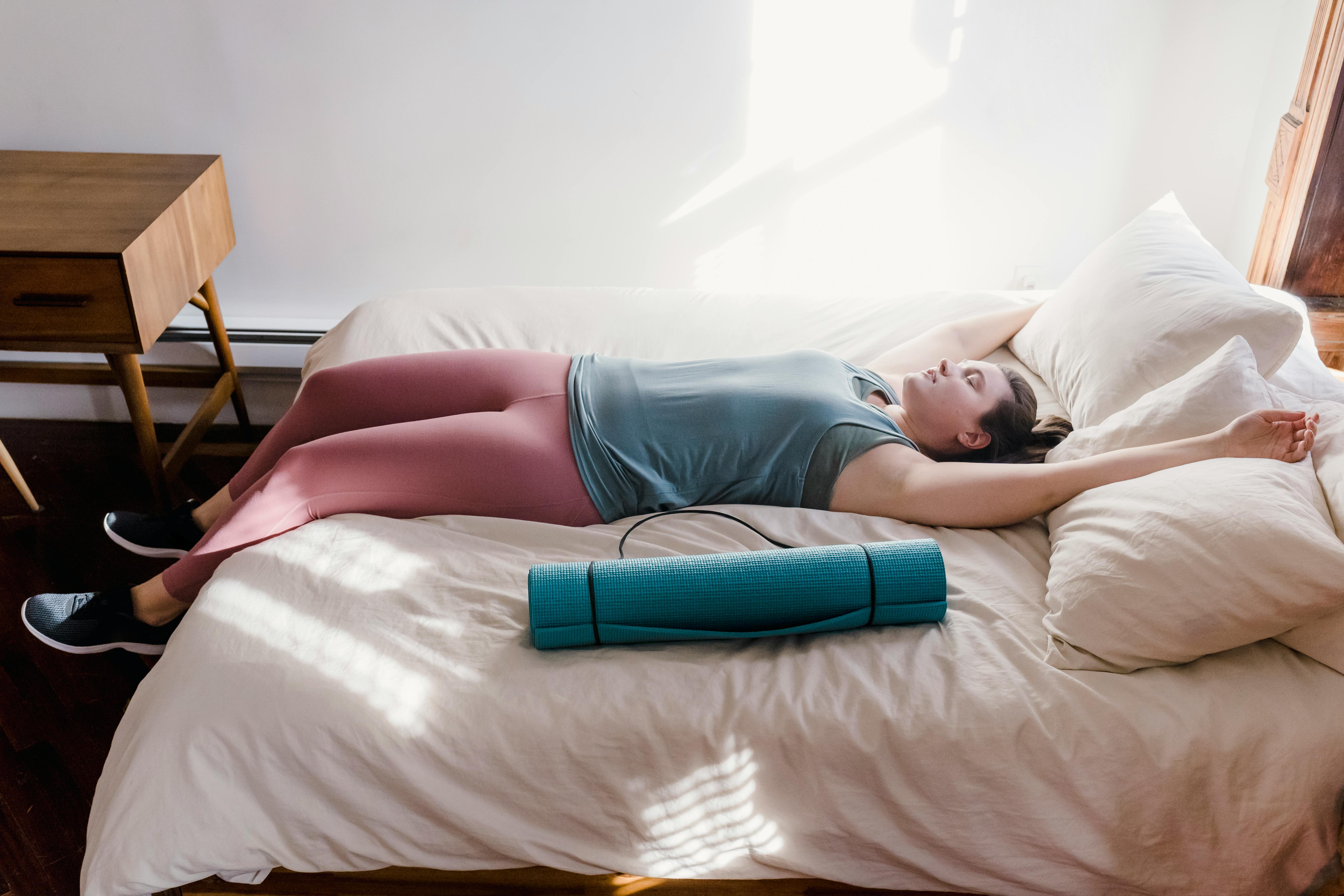 A woman in activewear lying on a bed with a yoga mat, enjoying sunlight and relaxation indoors.