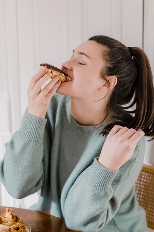 Happy female with eyes closed in sweater enjoying sweet chocolate doughnut at table at home