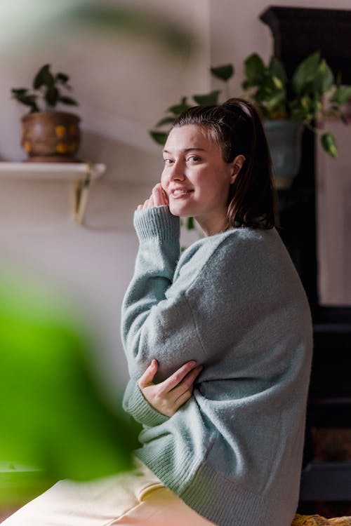 Side view of happy female in soft blue sweater smiling and looking at camera in apartment