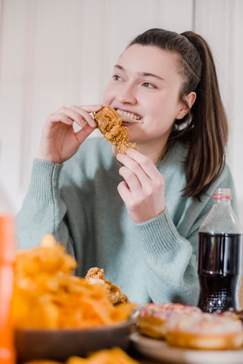 Happy female smiling and biting appetizing chicken while looking away in kitchen in daytime