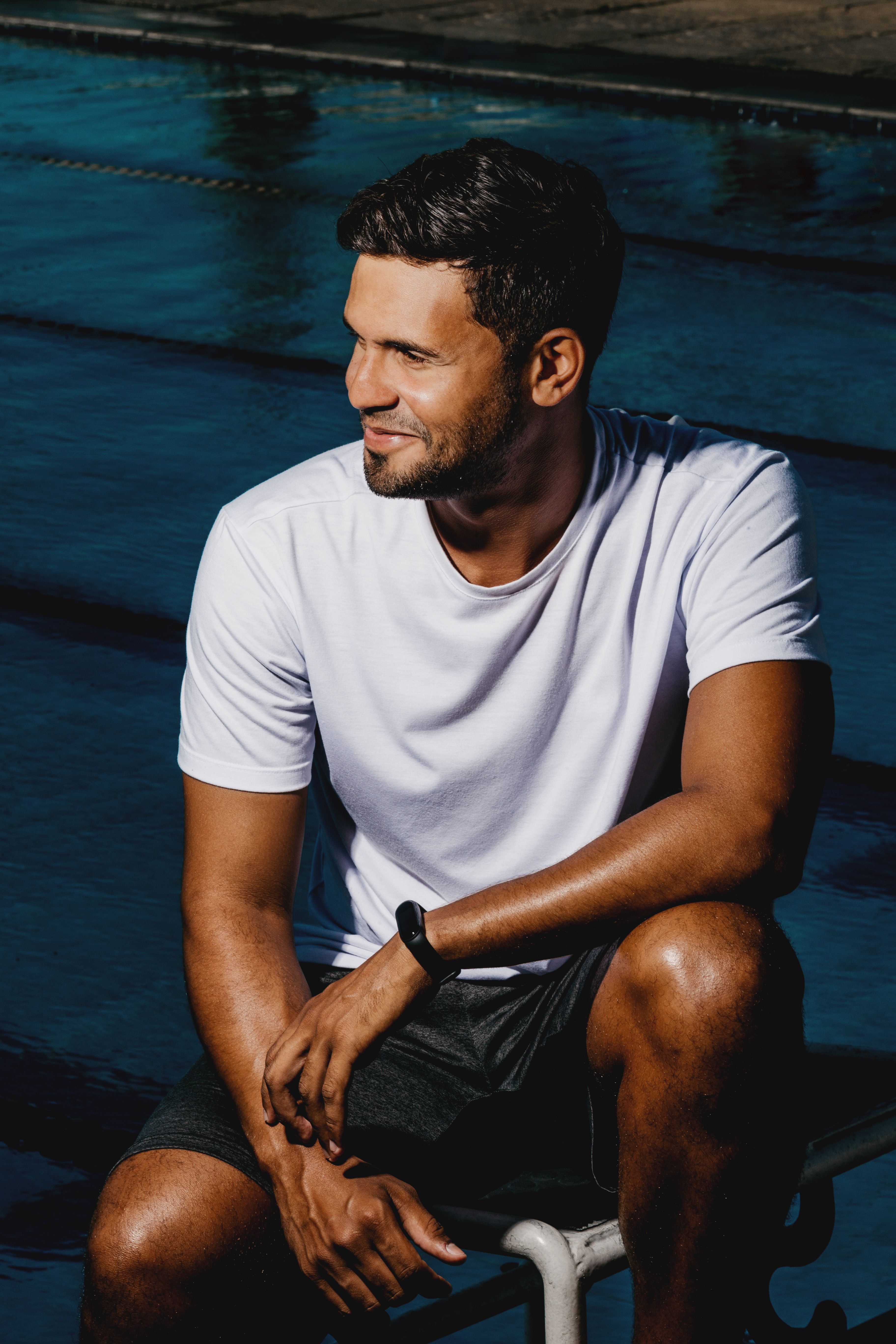 Positive male with beard wearing casual clothes sitting on haunches near swimming pool with clear water on sunny street in summer day