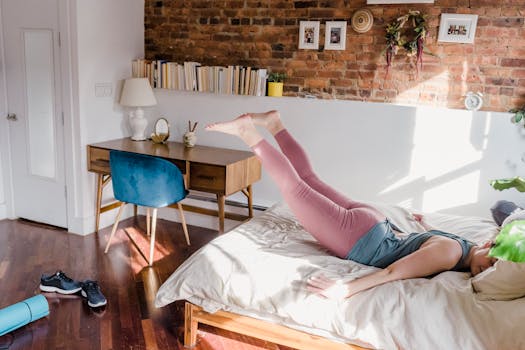 A woman in active wear exercises on a bed in a cozy sunlit bedroom with stylish decor.