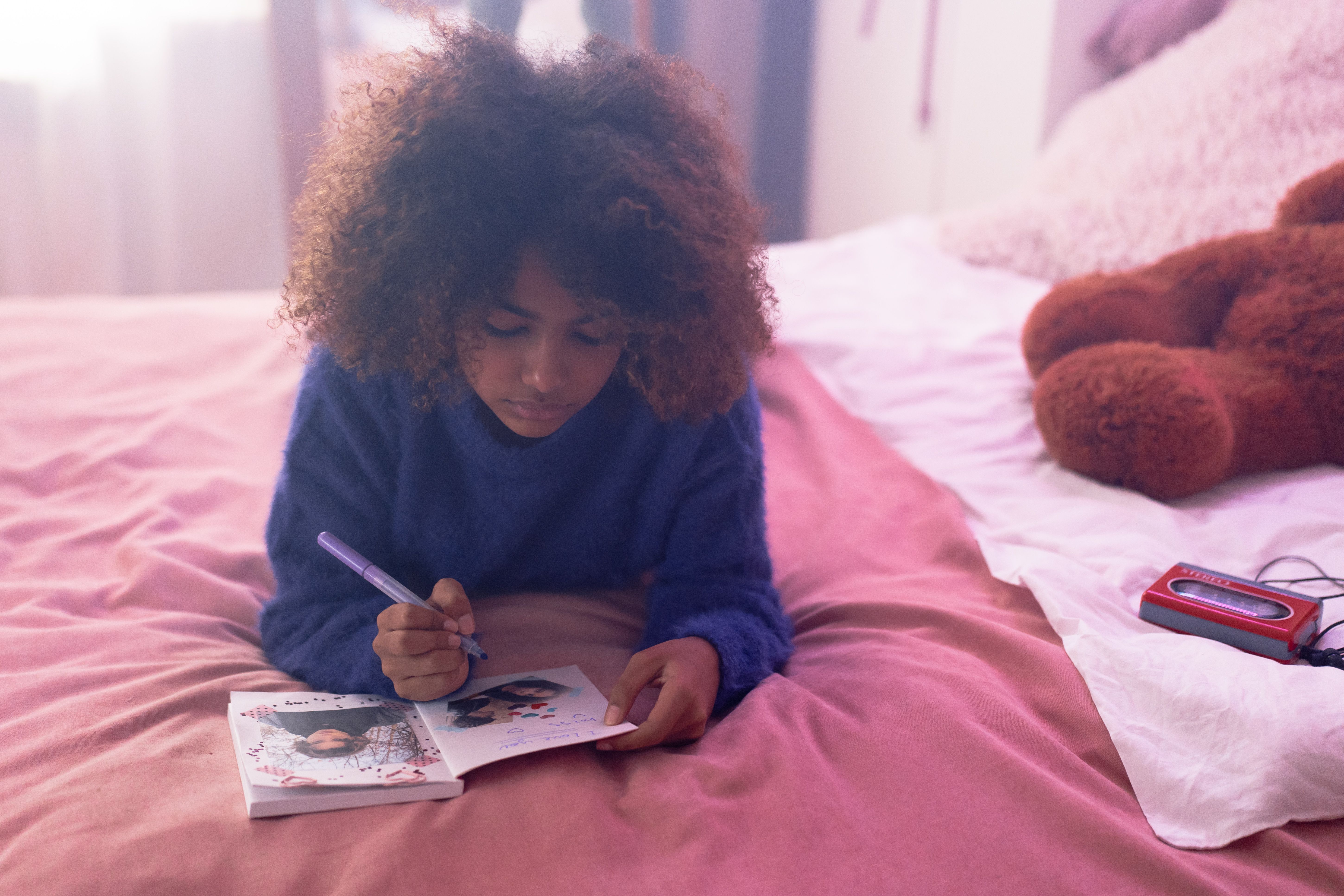 A young girl with afro hair writes in a journal while lying on a bed.