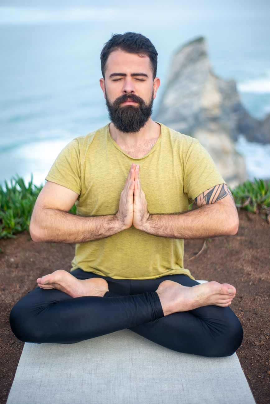 Bearded man meditating in lotus pose on yoga mat by the ocean cliff in Portugal.