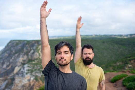 Two men standing on a cliff in Portugal, engaging in yoga exercises in a natural setting.