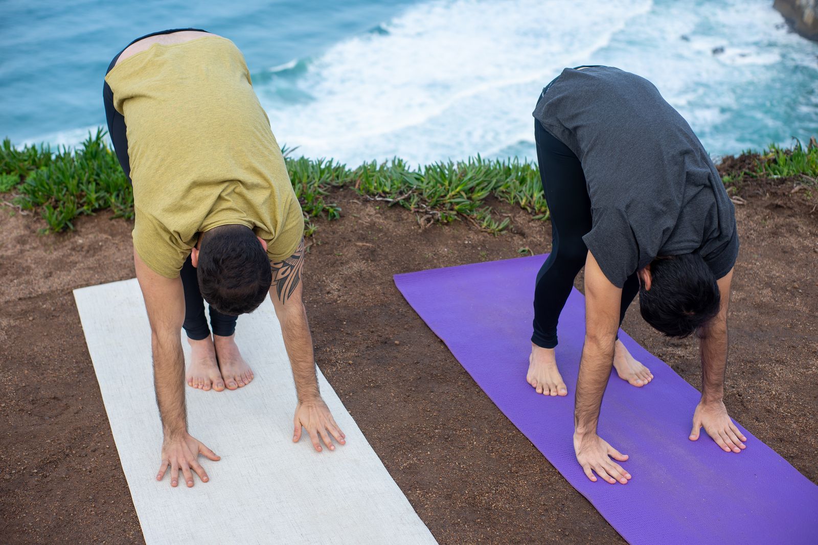 Two people practicing standing forward bend yoga pose outdoors by the sea
