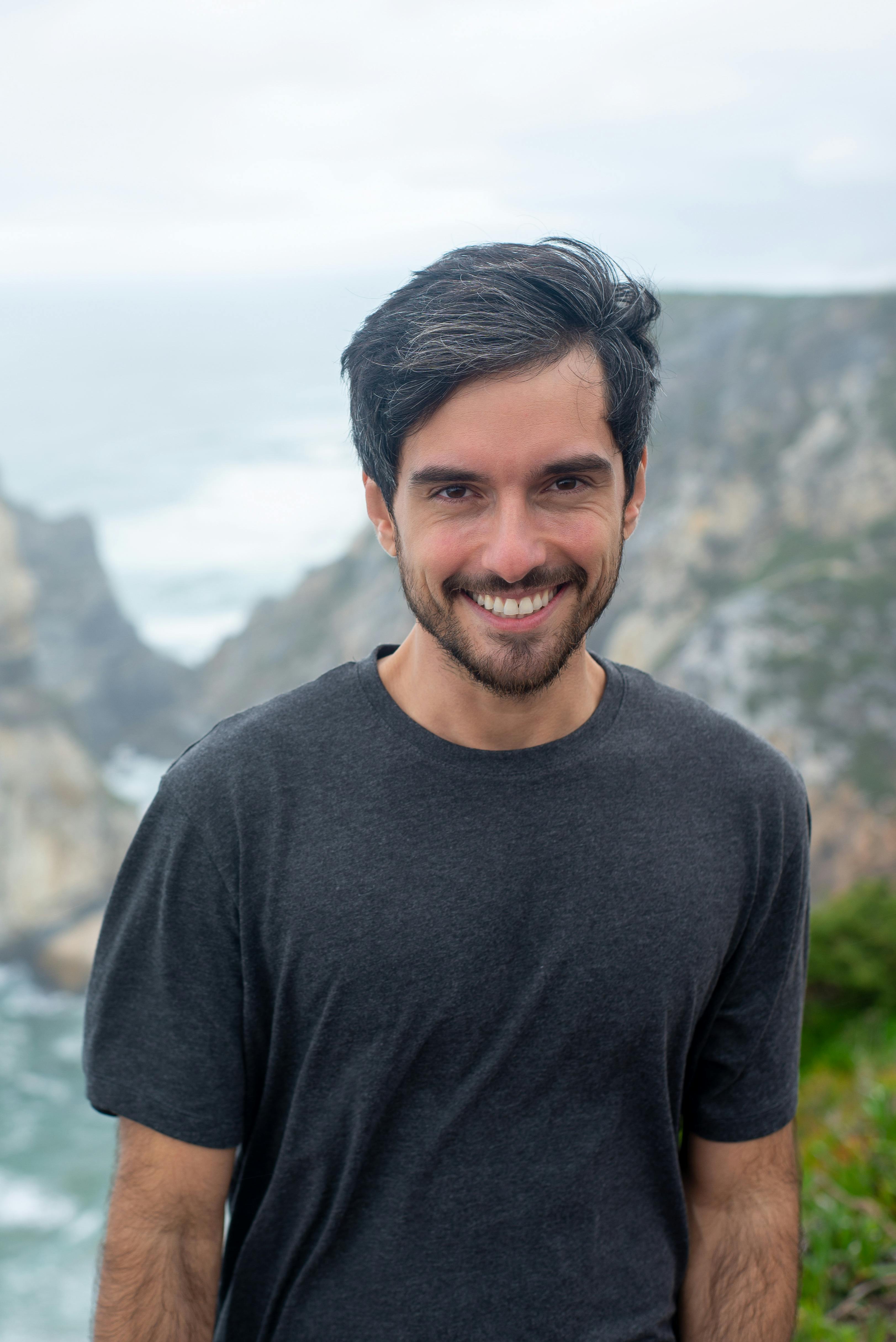 A smiling man in a black shirt poses in front of a scenic coastal cliff in Portugal.