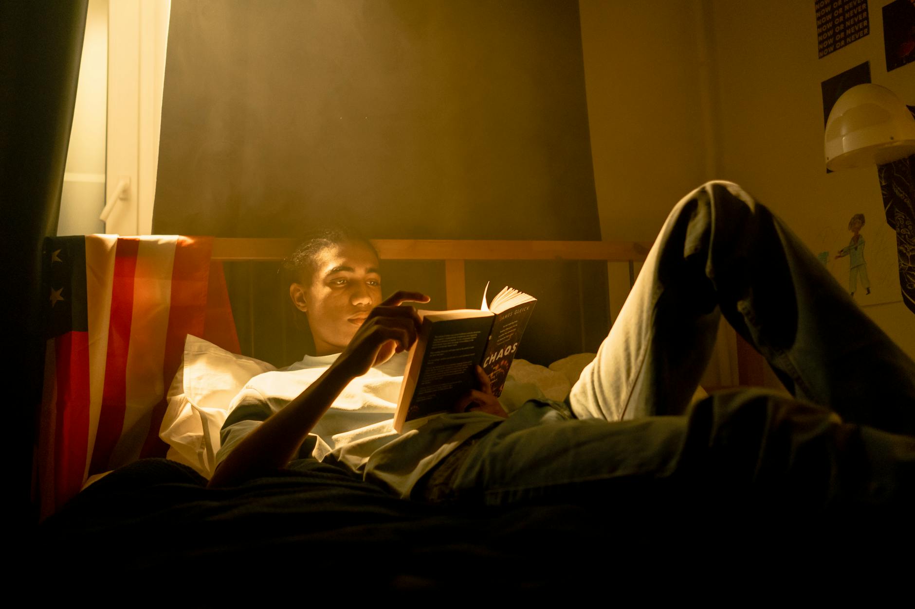 Teenager lying on bed reading a book under soft light in a cozy bedroom setting.