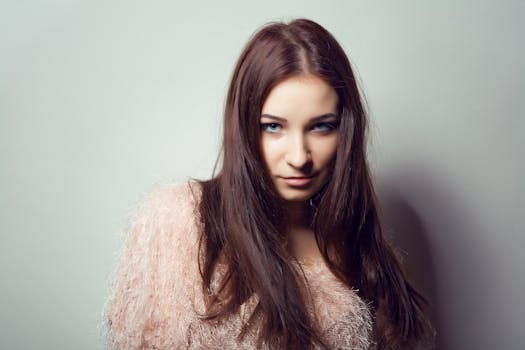 Close-up portrait of a young woman with long brown hair and a stylish outfit against a neutral background.