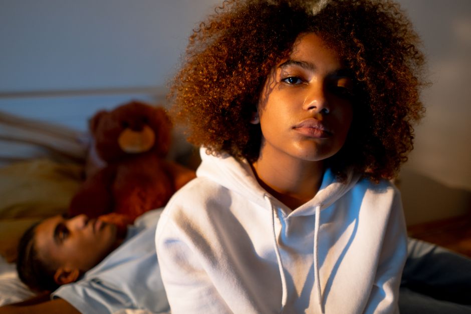 A young girl with curly hair wears a white hoodie, sitting pensively indoors.