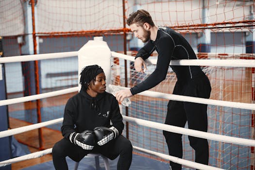 Two adults in a boxing ring with gloves, one seated and the other offering water.