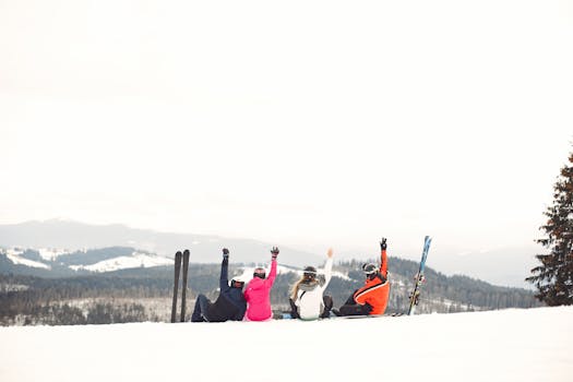 Four skiers sit on a snowy hill raising arms in joy, enjoying the scenic winter landscape.
