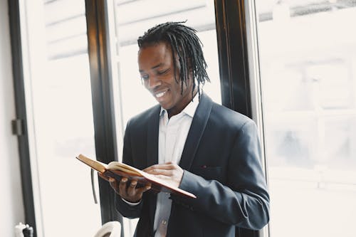 Man with Braided Hair Reading a Book by a Window and Smiling