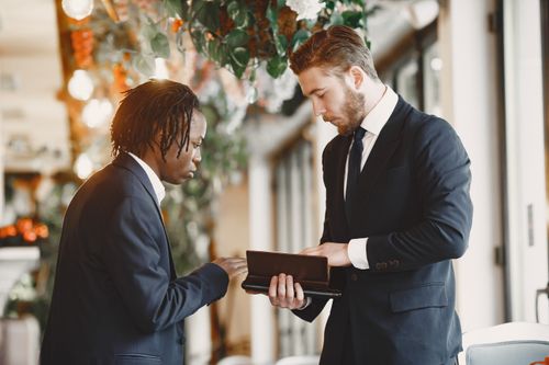 Free Two businessmen in suits discuss a document in a modern indoor setting with hanging plants. Stock Photo