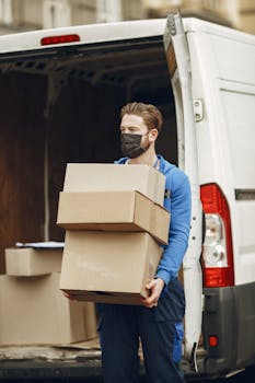Man wearing mask delivering cardboard boxes from van outdoors.