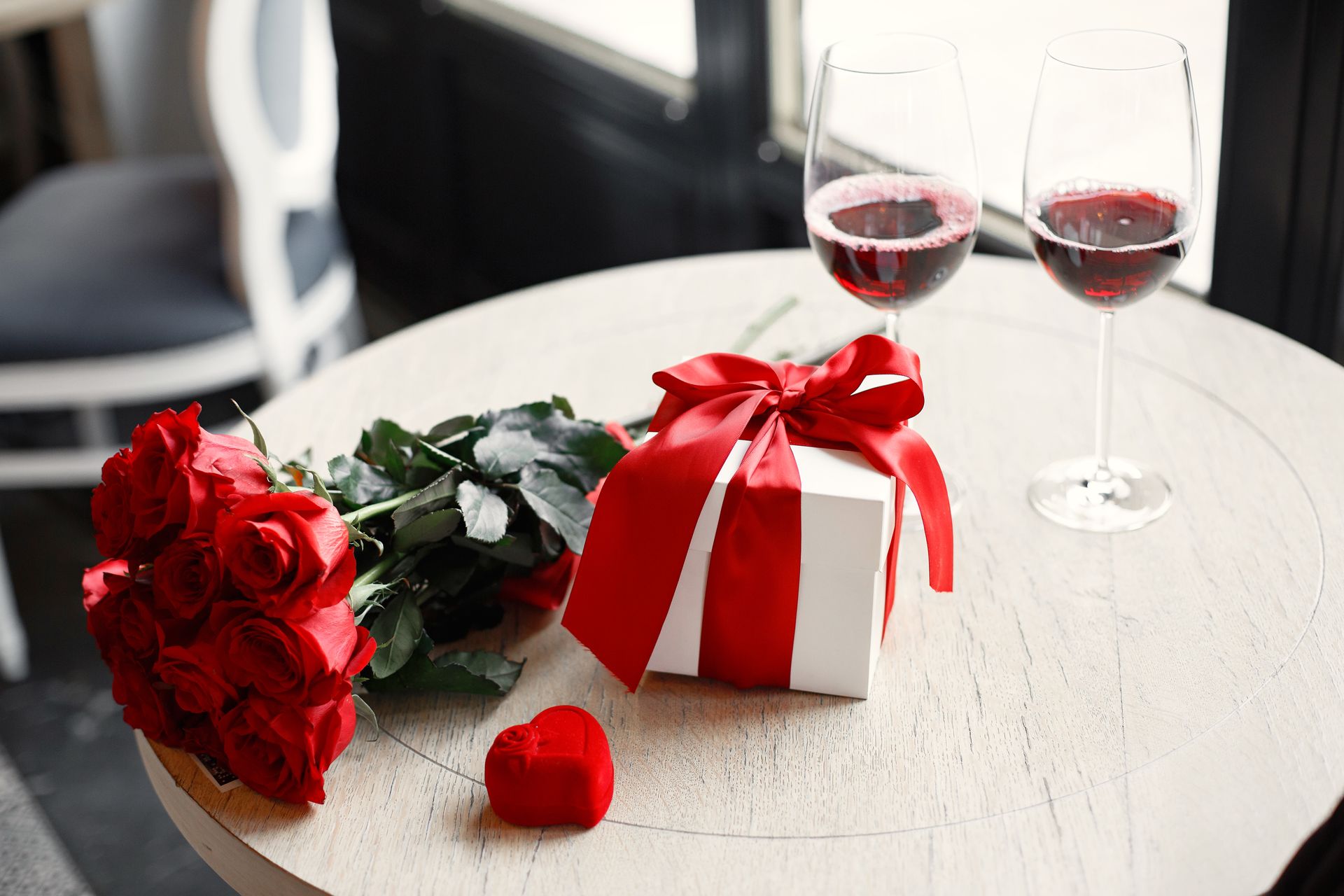 A romantic table setting with red roses, gift box, and red wine glasses, perfect for Valentine's Day celebration.