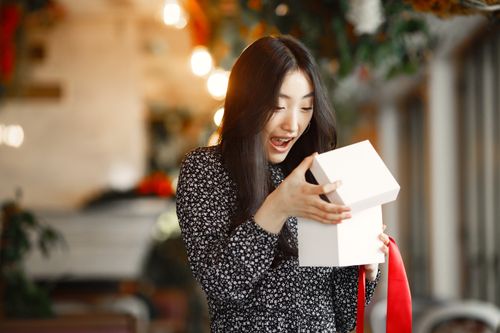 Free An Asian woman delightfully opens a gift box with a red ribbon indoors. Stock Photo