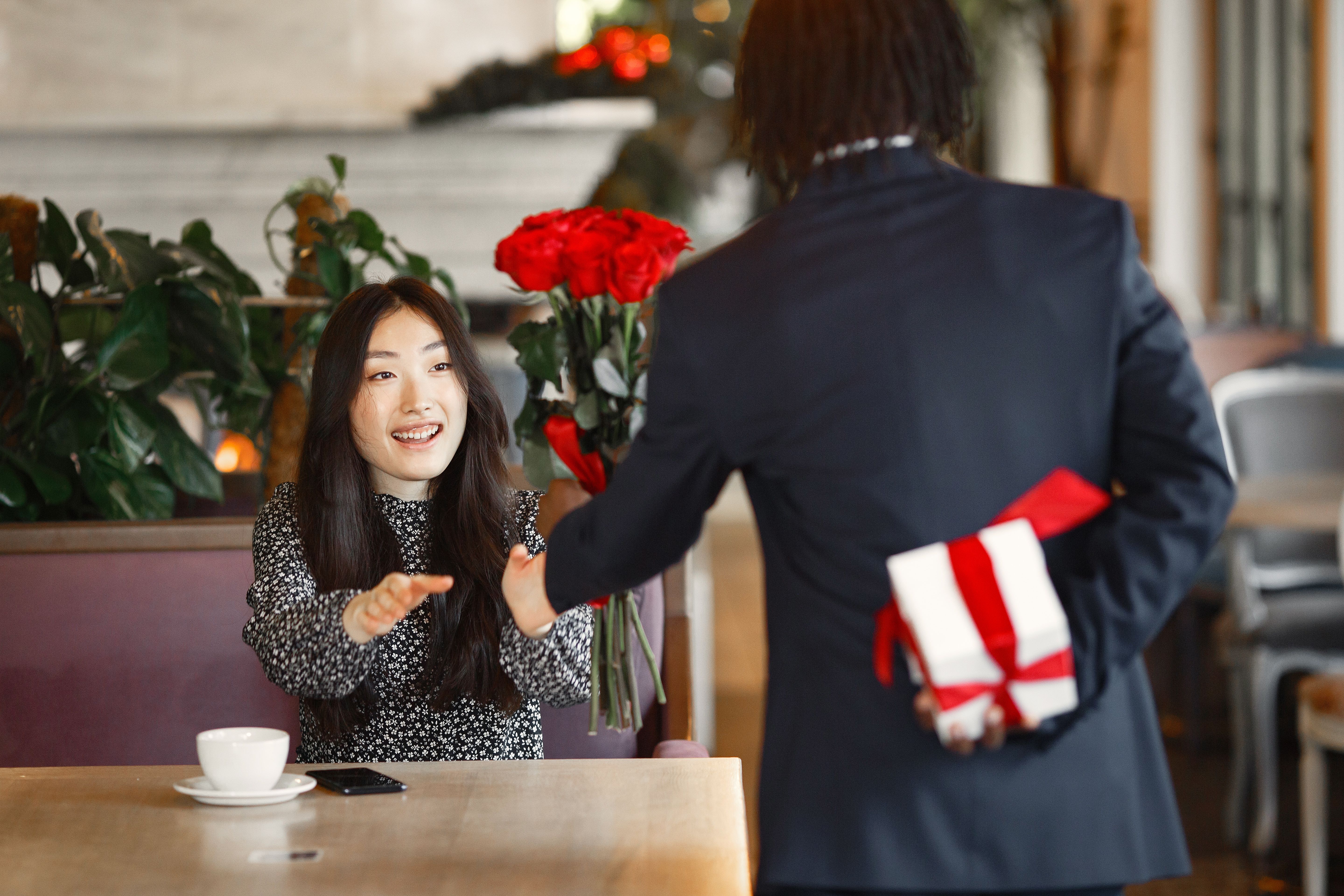 A man surprises a woman with red roses and a gift in a cozy cafe, capturing a moment of romance.