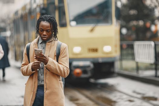A young man in a coat uses his smartphone while waiting for a tram in an urban setting.