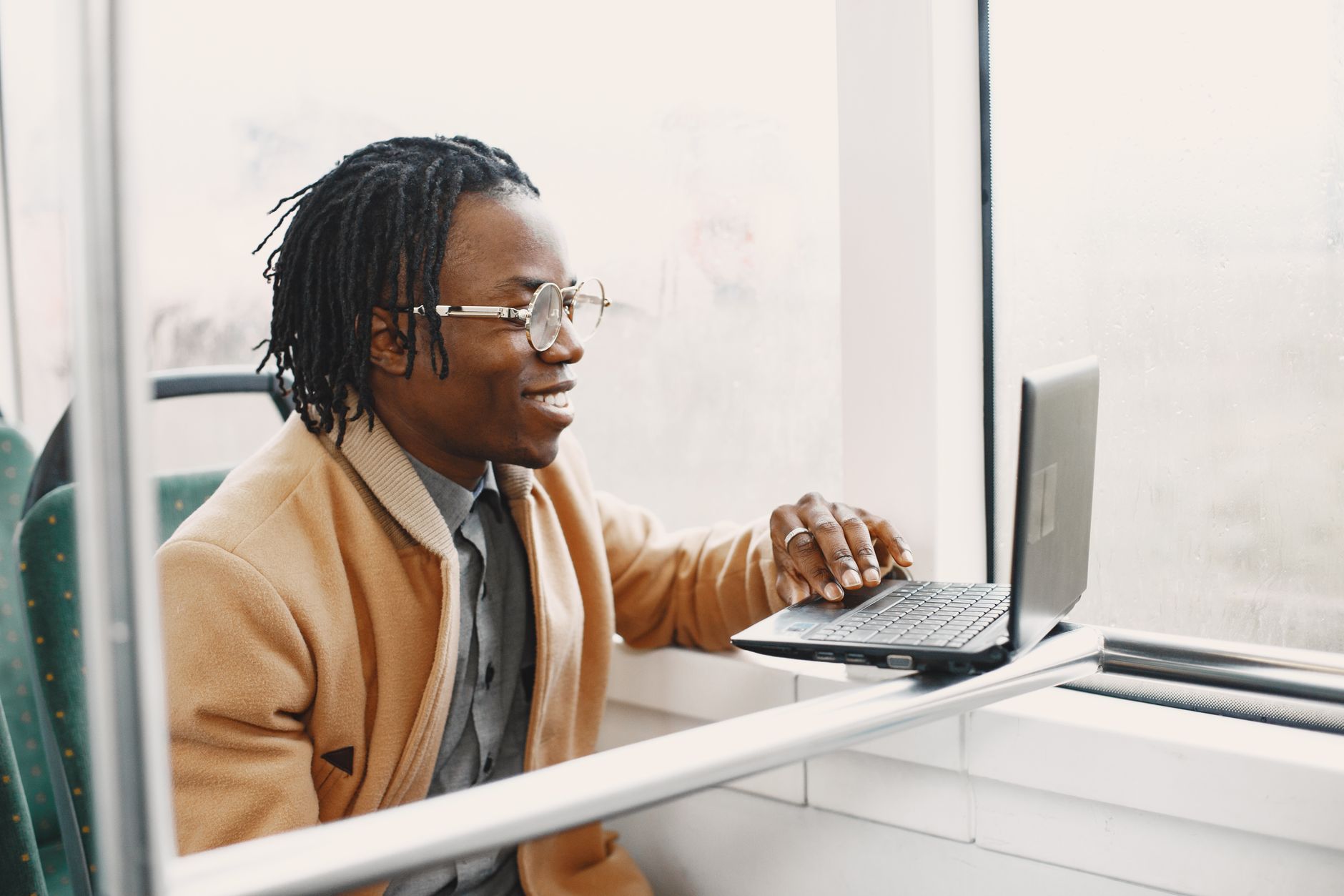 A stylish young man with dreadlocks smiles while working on a laptop during a commute.